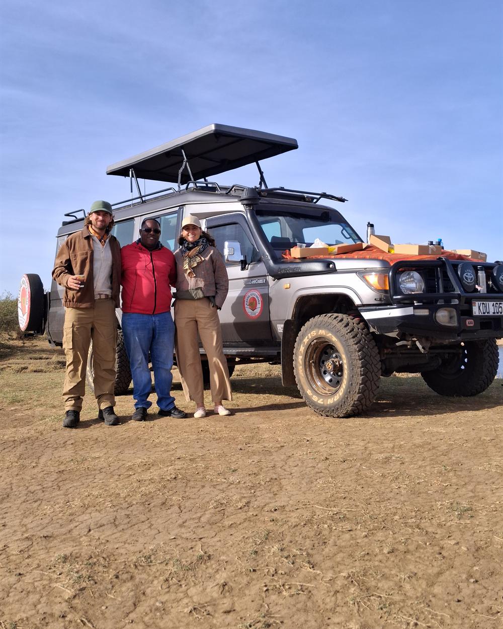 A driver guide in a red jacket with his arms around two guests beside a pop-up roof 4x4 safari vehicle, with a bush breakfast set out on the bonnet