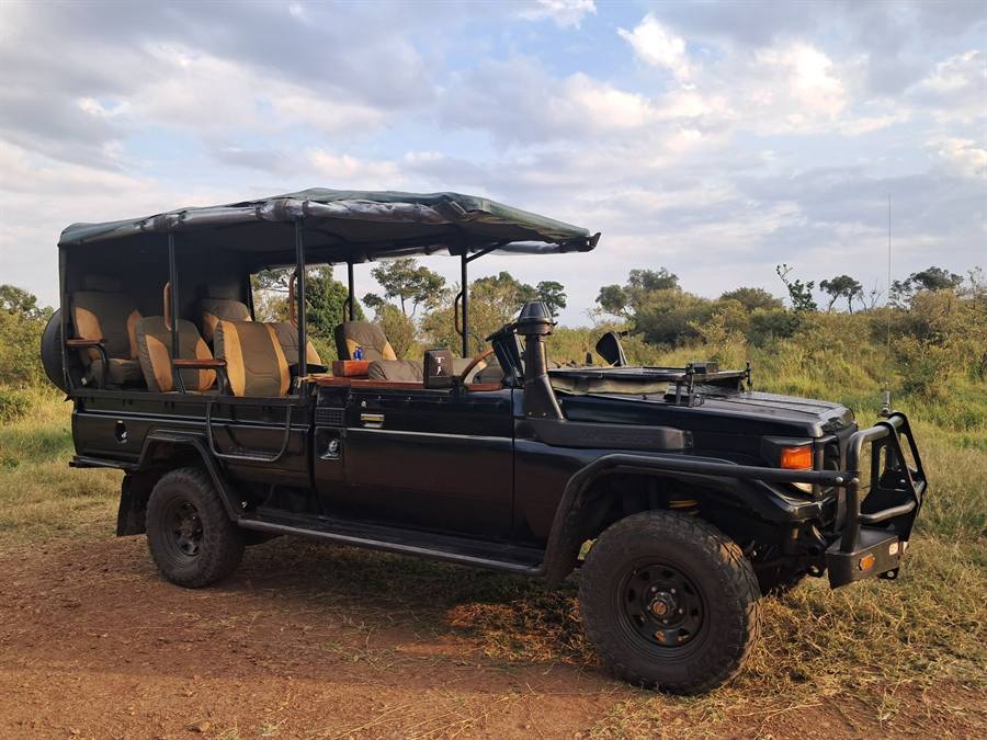 A black open-sided Land Cruiser game viewer with a canopy roof and tiered seats, parked on red earth in soft evening light