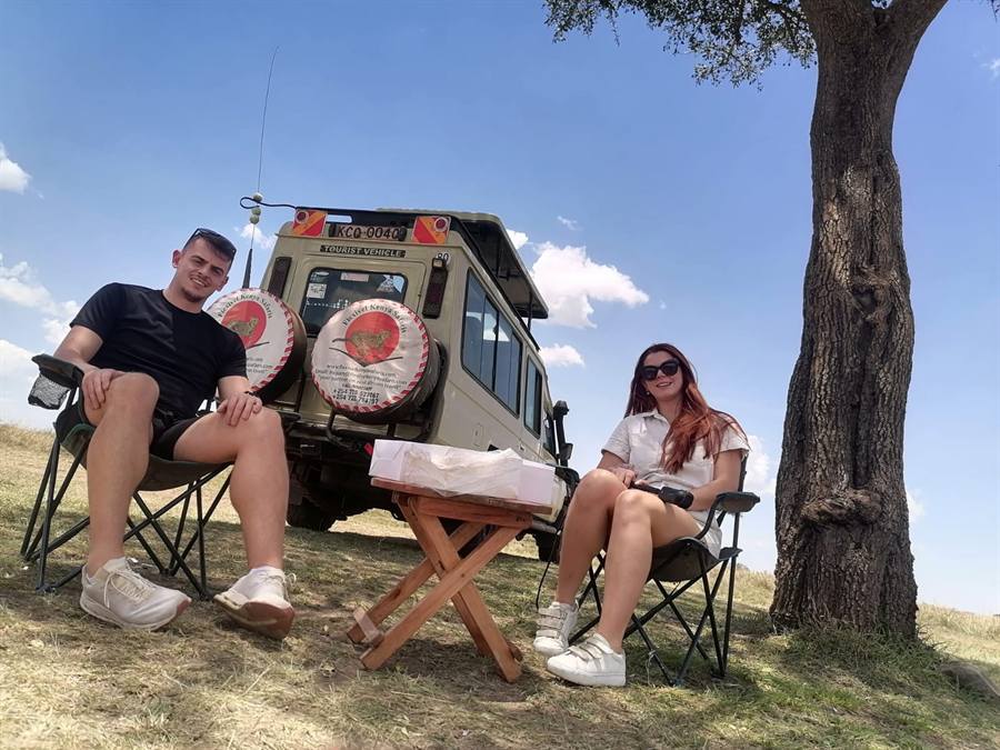 A couple relaxing in folding camp chairs beside a small table under a big tree, with a cream 4x4 safari vehicle behind them