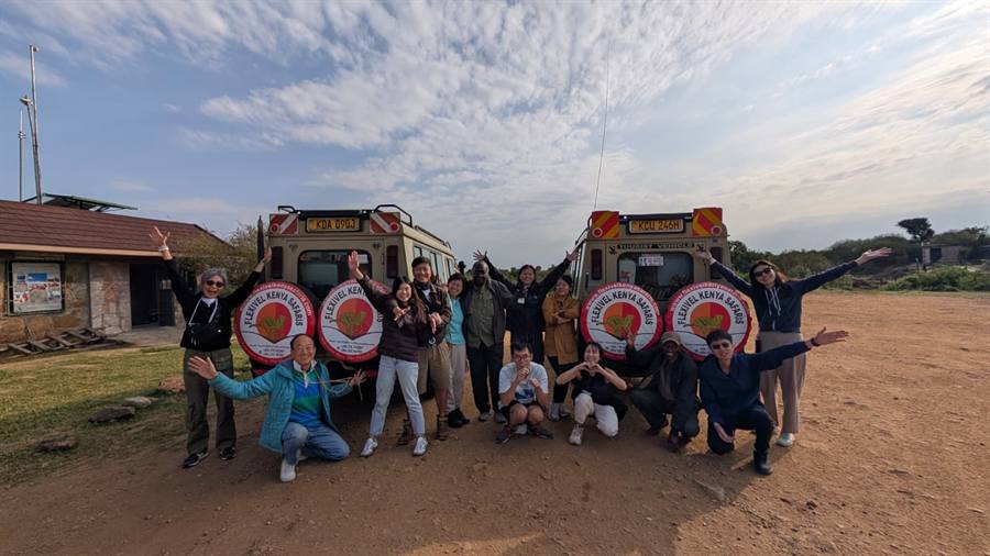 A large group of guests and guides posing with their arms up in front of two cream 4x4 safari vehicles with Flexivel Kenya Safaris wheel covers