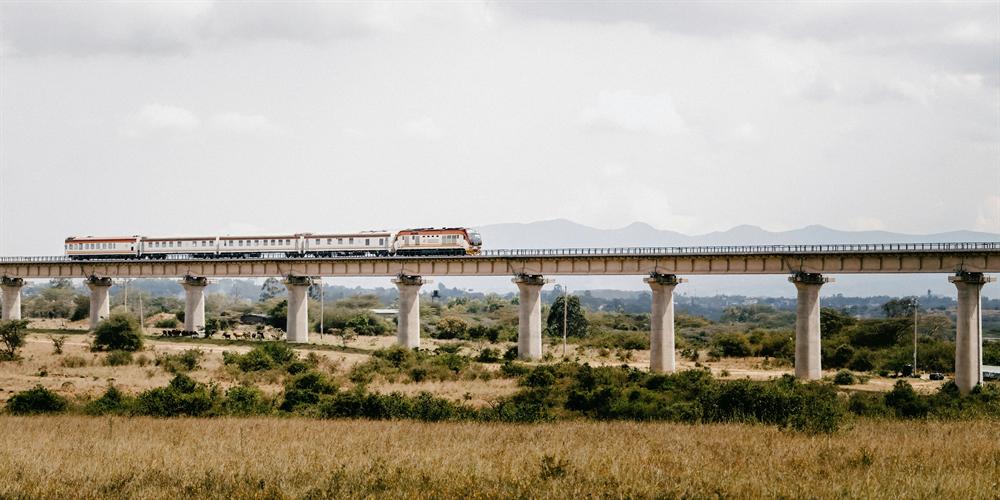 A Madaraka Express SGR passenger train crossing a long concrete viaduct on the plains of Kenya with hills behind