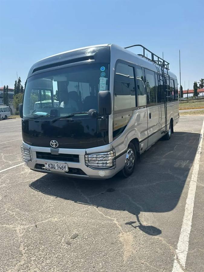 A silver and black Toyota Coaster bus with a roof rack, parked on a sunny car park