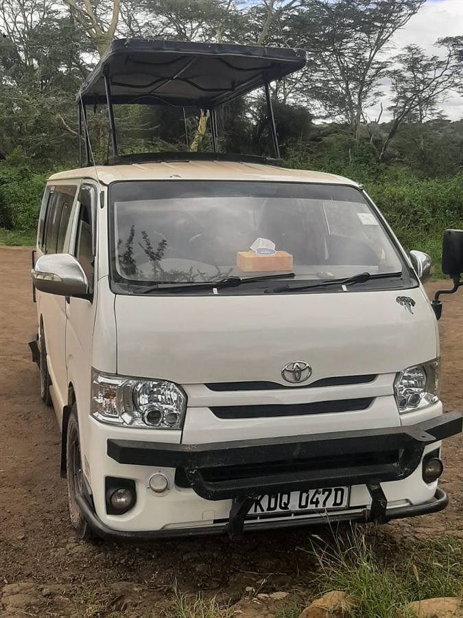 The front of a white Toyota safari van with its pop-up roof raised, parked under acacia trees