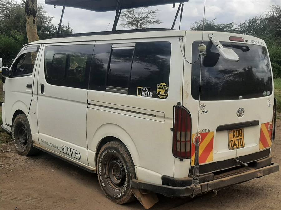 A white Toyota safari van with its pop-up roof raised, seen from the rear and side among acacia trees